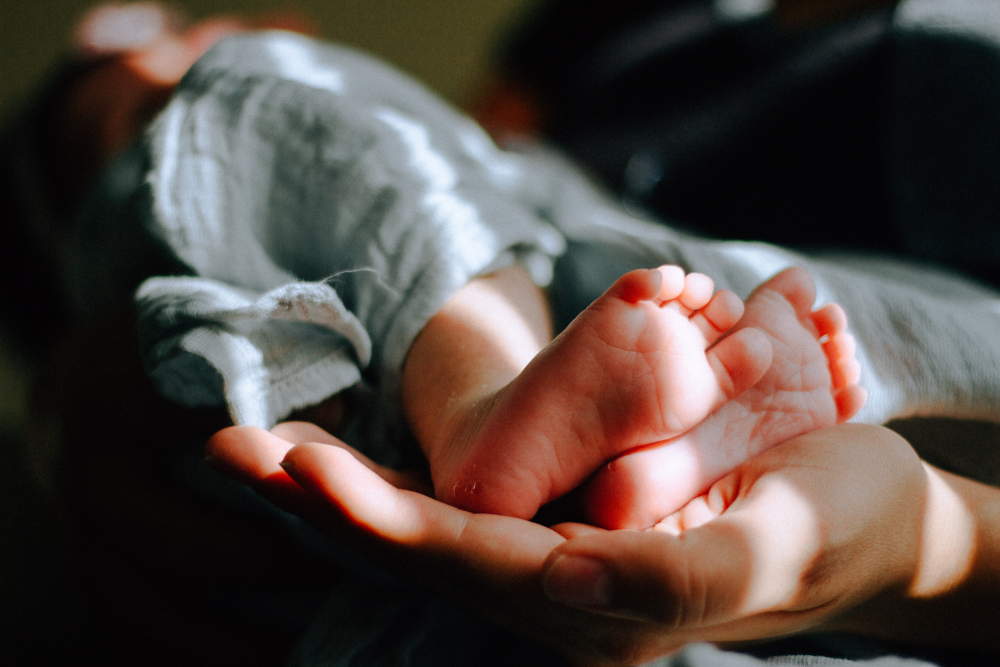 babyfuesschen-liegen-in-hand Eine Hand der Eltern hält zwei Babyfüsschen. Die Beine des Babys sind mit einem weißen Tuch bedeckt.