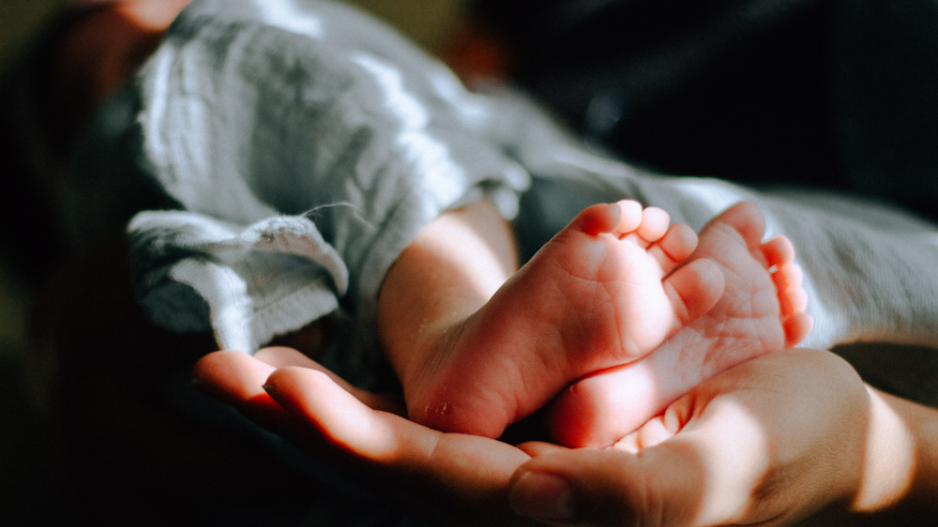 babyfuesschen-liegen-in-hand Eine Hand der Eltern hält zwei Babyfüsschen. Die Beine des Babys sind mit einem weißen Tuch bedeckt.