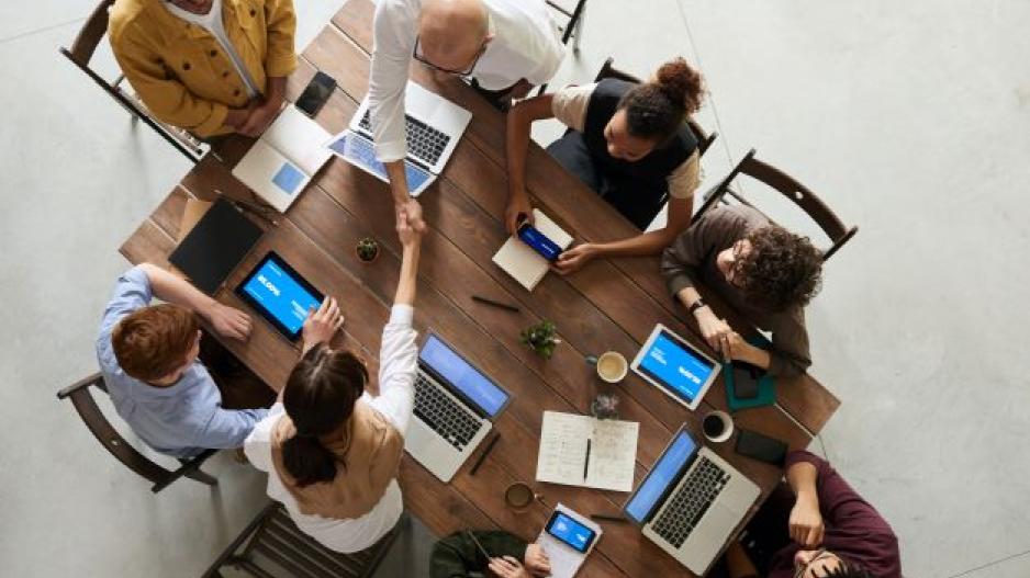 Eine Gruppe an Menschen unterschiedlichen Alters und Nationalität sitzt an einem Tisch zusammen. Alle arbeiten mit diversen Geräten am Tisch. Zwei Personen schütteln sich über den Tisch hinweg die Hände. Alle arbeiten gemeinsam. Kollaboration.