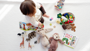 Baby sitzt auf weißer Decke und spielt mit einer Vielfalt an Spielsachen. Hier finden sich Stofftiere, Bücher, Stoffgemüse und kreative Spielideen für Kleinkinder.