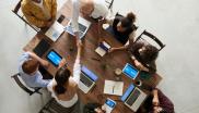 Eine Gruppe an Menschen unterschiedlichen Alters und Nationalität sitzt an einem Tisch zusammen. Alle arbeiten mit diversen Geräten am Tisch. Zwei Personen schütteln sich über den Tisch hinweg die Hände. Alle arbeiten gemeinsam. Kollaboration.