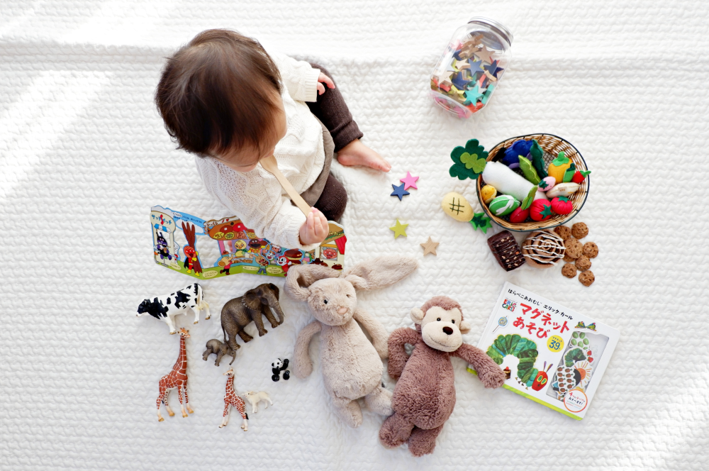 baby-beim-spielen Baby sitzt auf weißer Decke und spielt mit einer Vielfalt an Spielsachen. Hier finden sich Stofftiere, Bücher, Stoffgemüse und kreative Spielideen für Kleinkinder.