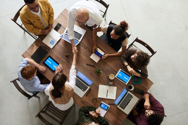 menschen-arbeiten-am-pc-zwei-schuetteln-haende.jpg Eine Gruppe an Menschen unterschiedlichen Alters und Nationalität sitzt an einem Tisch zusammen. Alle arbeiten mit diversen Geräten am Tisch. Zwei Personen schütteln sich über den Tisch hinweg die Hände. Alle arbeiten gemeinsam. Kollaboration.